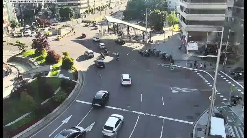 A sunny day shows a city intersection with numerous cars, motorcycles, and pedestrians, centered around a modern, open-air bus shelter.