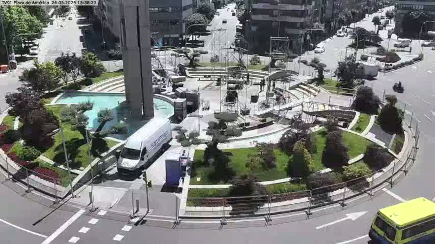 A sunny day reveals a city plaza under construction, featuring a central fountain, landscaping, and various vehicles parked around the perimeter.