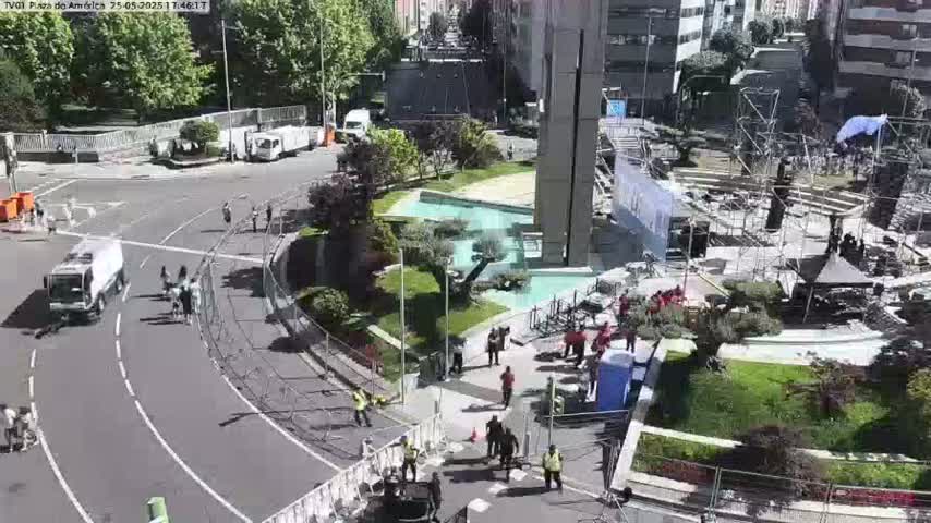 A sunny city square shows a street scene with vehicles, pedestrians, and preparations for an event, including stage scaffolding and a water feature.