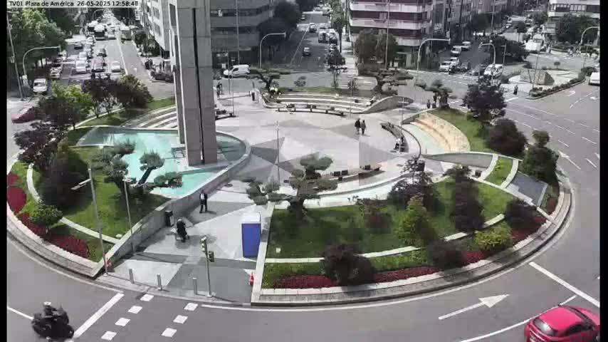 A circular plaza with a water feature and landscaping is surrounded by roads and buildings on a sunny day.