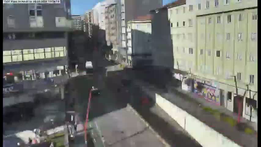 A high-angle, sunny-day view of a city street scene showing multi-story buildings, a roadway, and some pedestrians.