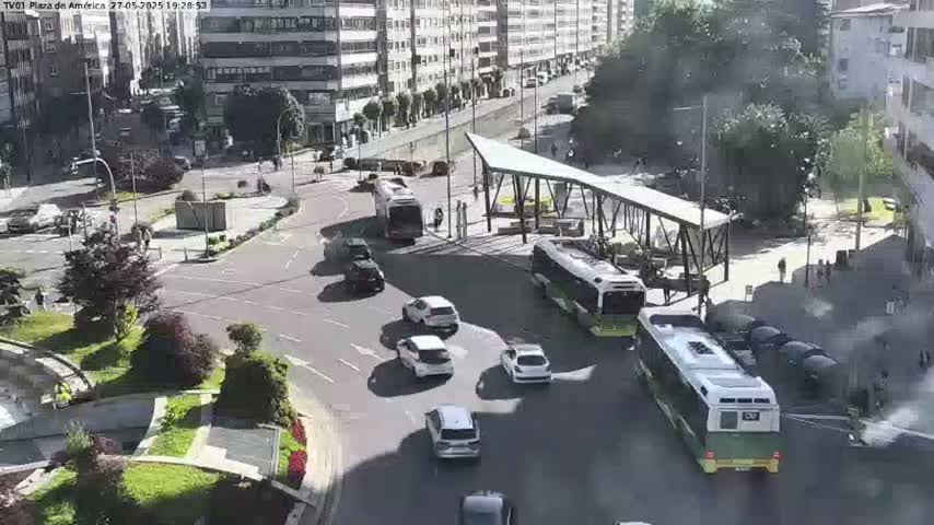 A sunny day reveals a plaza with several cars and buses navigating a roundabout, surrounded by buildings and landscaping.