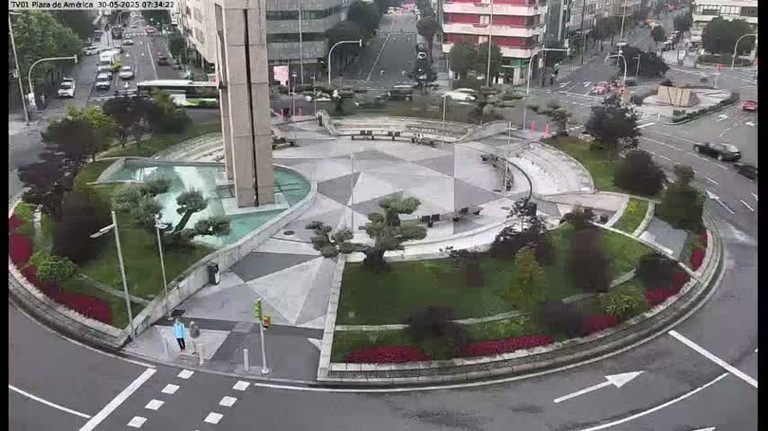 An aerial view of a landscaped traffic circle with a geometric pattern in the center, surrounded by low shrubs and trees, on a slightly overcast day.