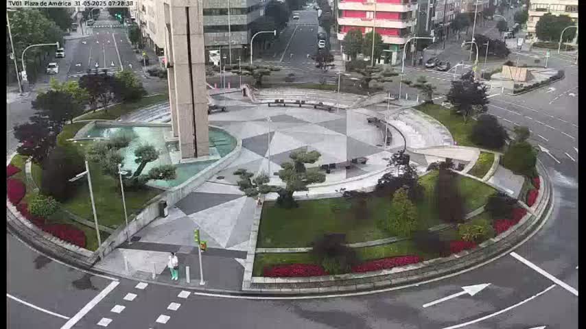 An aerial view shows a circular plaza with geometric landscaping, a water feature, and surrounding buildings on a wet, overcast day.