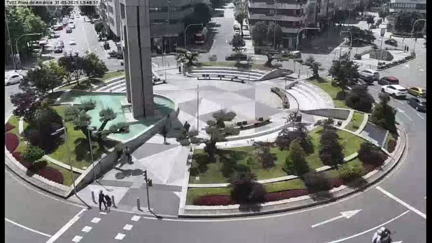 A sunny aerial view shows a circular plaza with landscaping, a water feature, and several people walking, surrounded by a roadway with cars driving.