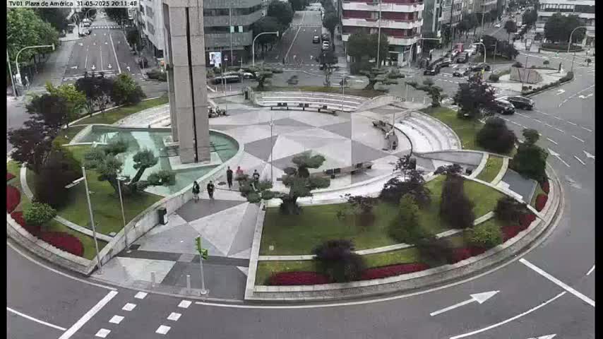 A paved, circular plaza with landscaping and a fountain is surrounded by roads with cars and buildings visible in the background on an overcast day.