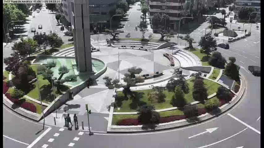 A sunny day reveals a circular plaza with a fountain, landscaped areas, and seating, surrounded by a roadway with sparse traffic.