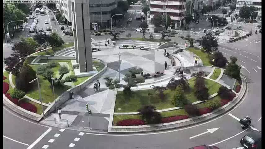 An aerial view of a circular plaza with landscaped gardens and walkways, surrounded by roads with cars and a few pedestrians, on a partly cloudy day.