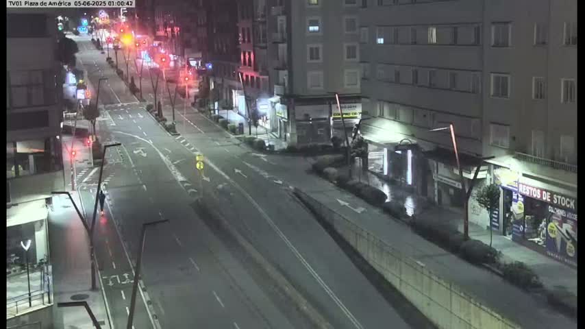 A mostly empty, city street at night is seen from an elevated vantage point, with a few vehicles sparsely distributed along its lanes and buildings lining the street.