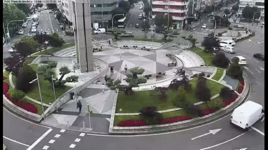 A circular plaza with landscaped gardens and a star-shaped paved center is surrounded by roads with cars and a white van turning the corner, on an overcast day.