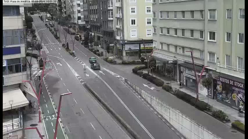 A mostly empty city street with two cars driving on it, flanked by multi-story buildings under a cloudy sky.