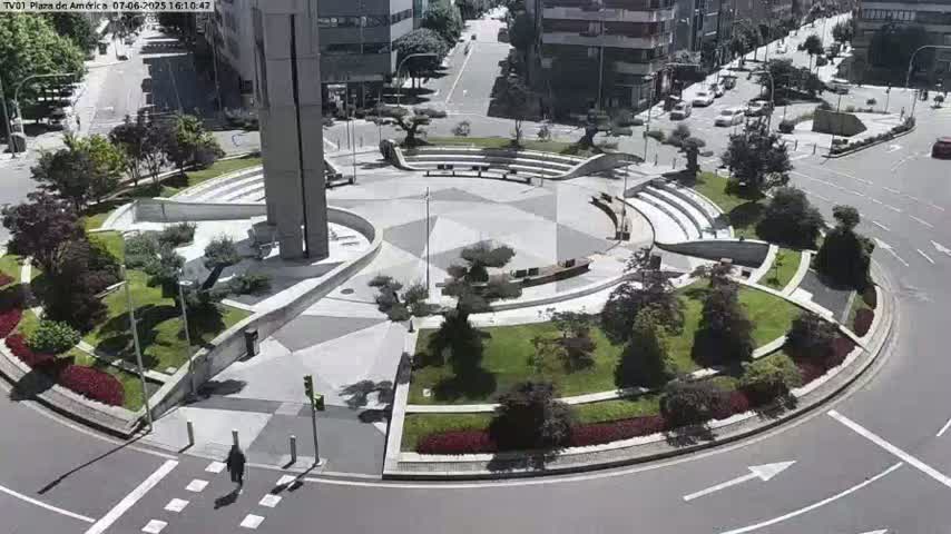 A circular plaza with landscaping, seating areas, and a central monument is surrounded by a roadway with sparse traffic under sunny conditions.