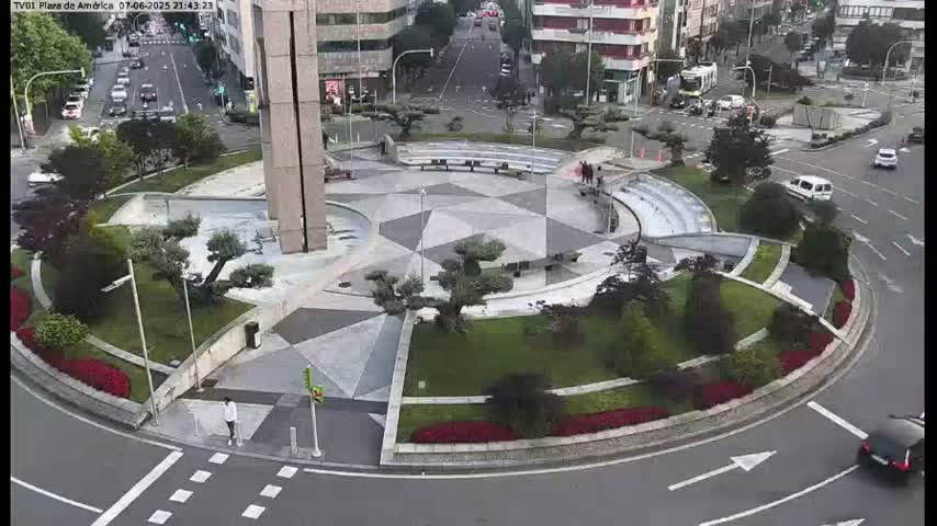 A circular plaza with geometric landscaping and a tall monument in the center is surrounded by roads with cars and buildings in the background under a clear sky.