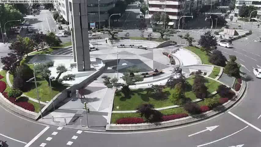 A sunny day reveals a landscaped traffic circle with a geometrically patterned plaza in the center, surrounded by green spaces and buildings.
