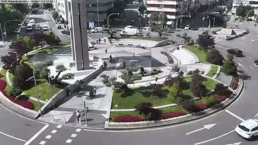 A sunny day reveals a landscaped roundabout with pedestrians and vehicles, surrounded by buildings.