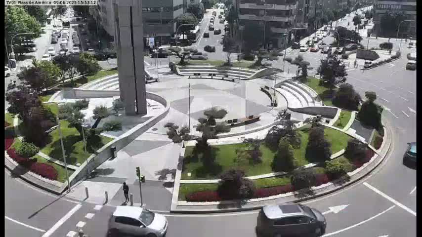 A circular plaza with landscaped gardens and seating areas is surrounded by a roadway with cars, under a sunny sky.