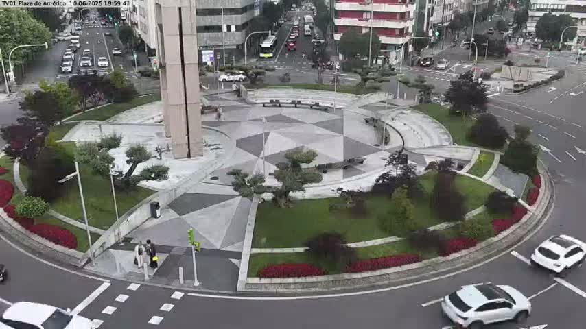 An aerial view of a city roundabout with a geometrically designed center, surrounded by landscaped greenery, under an overcast sky.
