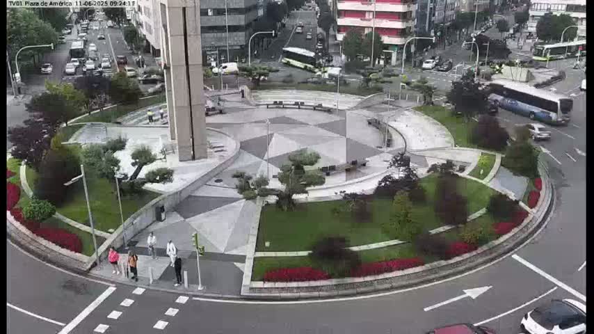 A circular plaza with geometric landscaping, small trees, and a few pedestrians is situated in the center of a roundabout surrounded by city streets and buildings, under an overcast sky.
