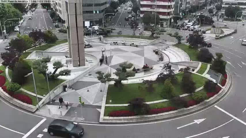 A circular plaza with landscaped gardens and a few pedestrians is surrounded by a roadway on a partly cloudy day.