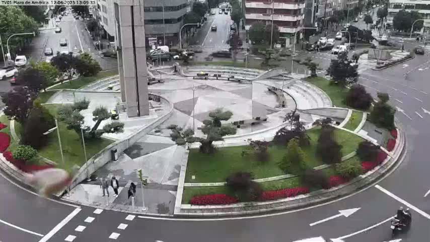 A circular plaza with landscaped gardens and a central paved area is surrounded by roads with cars and a person on a motorcycle; it is a cloudy day.