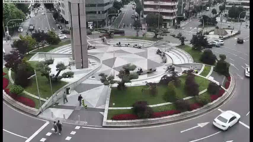A paved, geometric plaza with landscaped areas and a surrounding roadway, with several people walking and a few cars driving, on an overcast day.