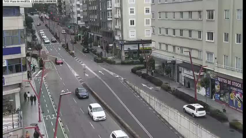 A city street scene on an overcast day shows several cars driving on a multi-lane road, with pedestrians walking on a sidewalk alongside a bike lane, near multi-story buildings.