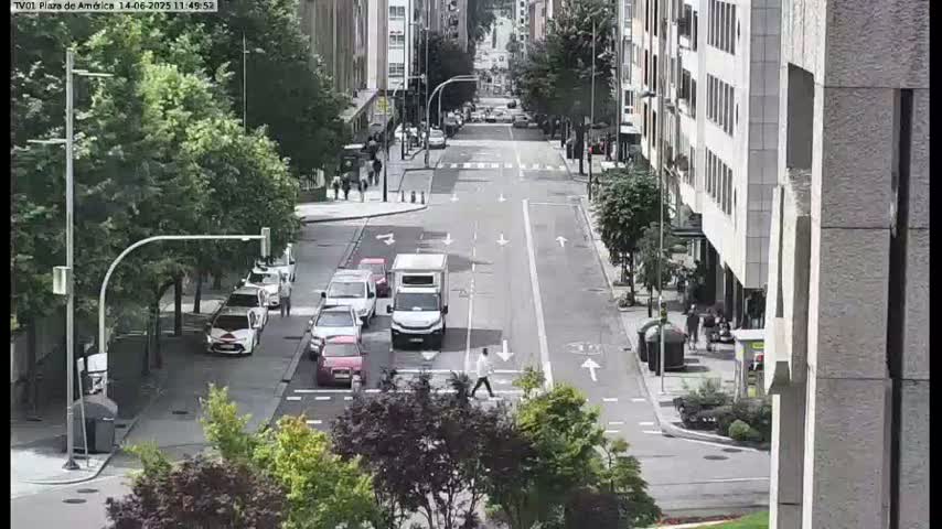 A city street scene on a partly cloudy day shows several cars stopped at a crosswalk with a pedestrian crossing the street, flanked by tall buildings and lush green trees.