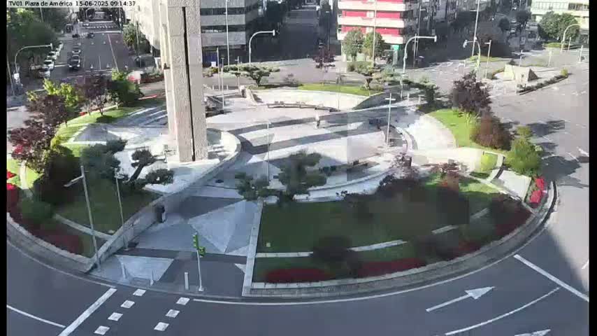 A sunny aerial view of a roundabout landscaped with green grass, trees, and flowerbeds, with a modern building and a few pedestrians visible.