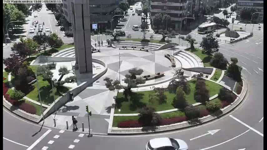 A sunny day shows a circular plaza with a geometrically designed landscaped center, surrounded by a roadway with cars and pedestrians.