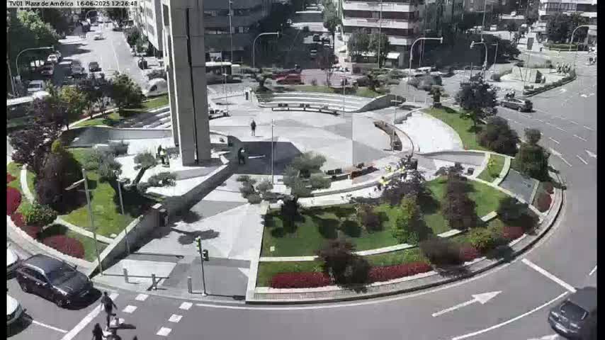 A high-angle view of a plaza with a circular roadway, landscaped areas, and several pedestrians, on a sunny day.