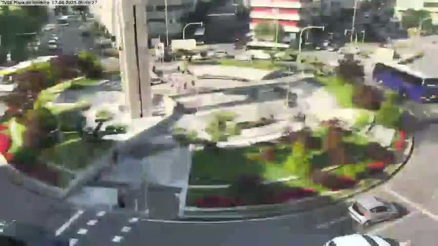 A sunny aerial view of a roundabout with lush landscaping, several cars, and a bus in the surrounding streets.