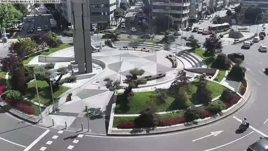 A landscaped roundabout with a central plaza, surrounded by roads with cars and a person on a scooter, on a sunny day.