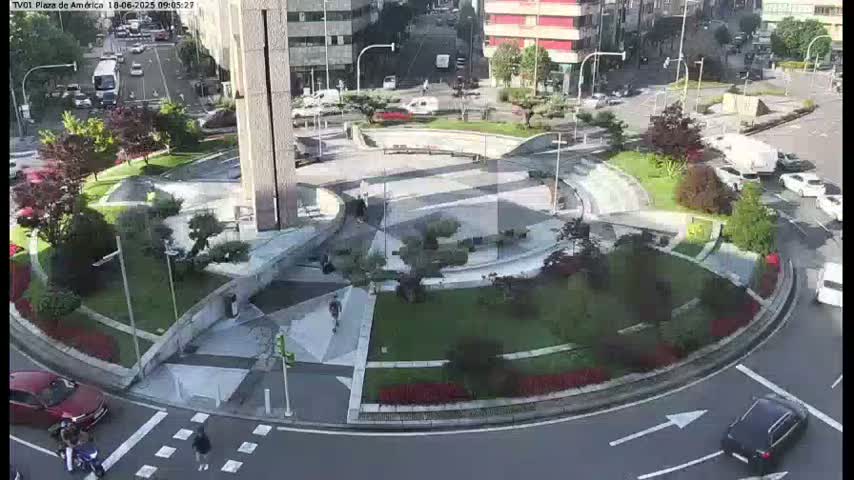 A paved roundabout with landscaped areas and a central plaza is surrounded by city streets with cars and pedestrians; the weather appears sunny.