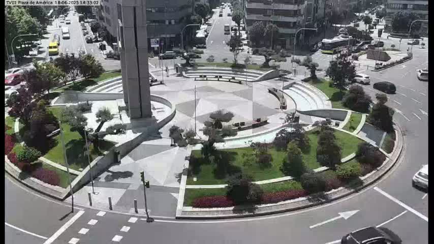 A sunny day reveals a circular plaza with landscaped gardens, seating areas, and a tall building, surrounded by roads with cars.
