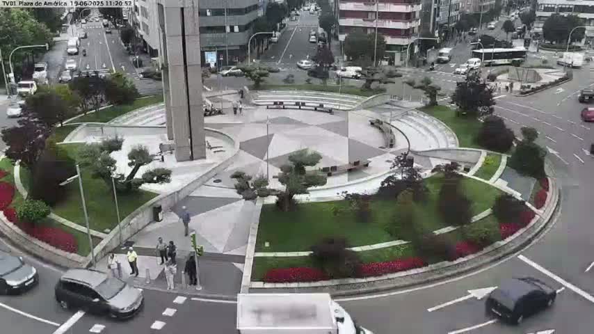 A roundabout in a city center, landscaped with shrubs and flowers, shows several cars driving around it on a partly cloudy day.