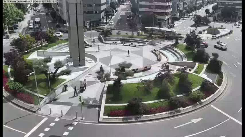 An aerial view of a circular plaza with landscaped gardens and seating areas, surrounded by roads with cars, under a sunny sky.