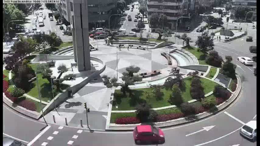 A circular plaza with landscaped gardens and seating areas is surrounded by a roadway with several cars, under a sunny sky.