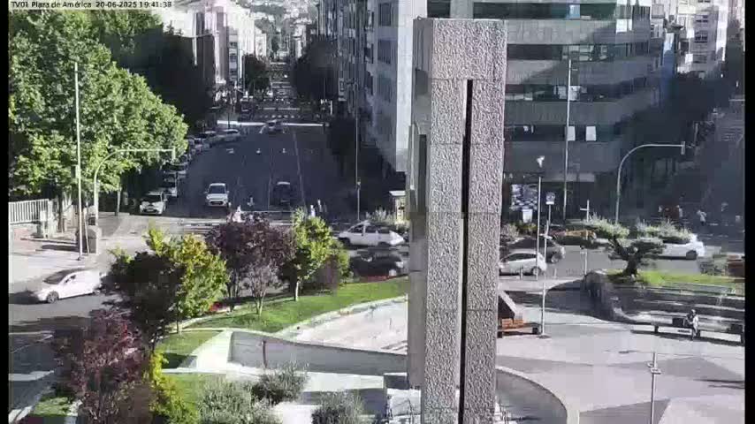 A sunny city square with a tall, grey stone monument in the center, surrounded by trees, parked cars, and a few pedestrians, with some traffic visible on nearby streets.
