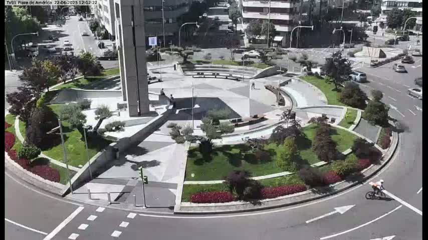 A circular plaza with landscaped gardens and a paved central area, surrounded by roads with cars and a cyclist, under sunny conditions.