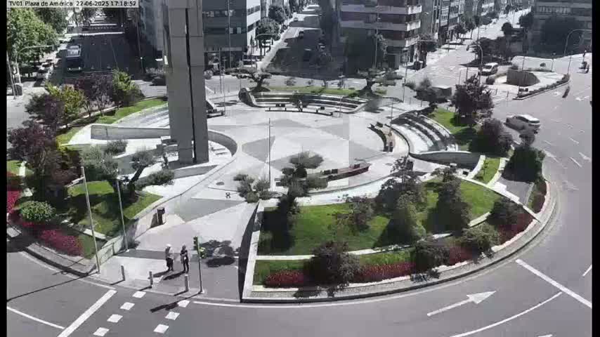 A sunny day reveals a circular plaza with landscaped gardens, seating areas, and a central paved area, surrounded by roadways and buildings.