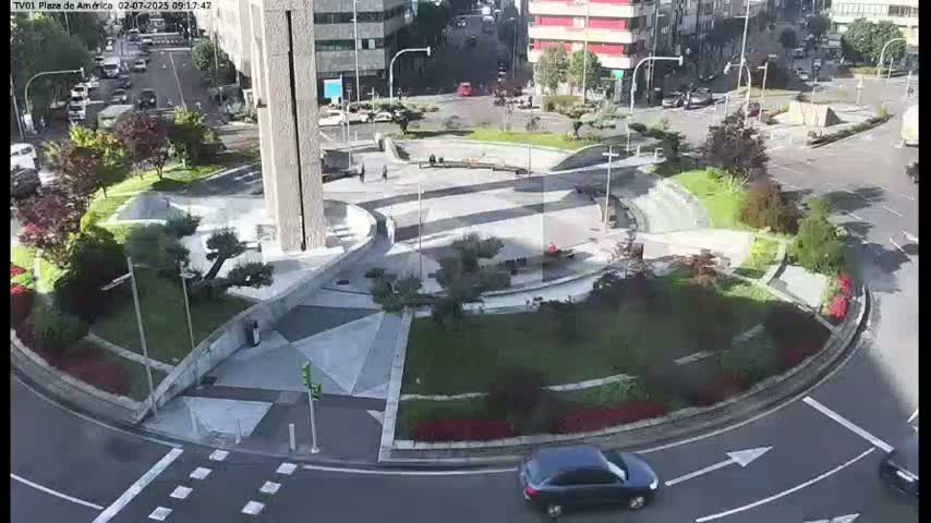 A circular plaza with landscaped gardens and a tall building in the background is viewed from above on a partly sunny day.