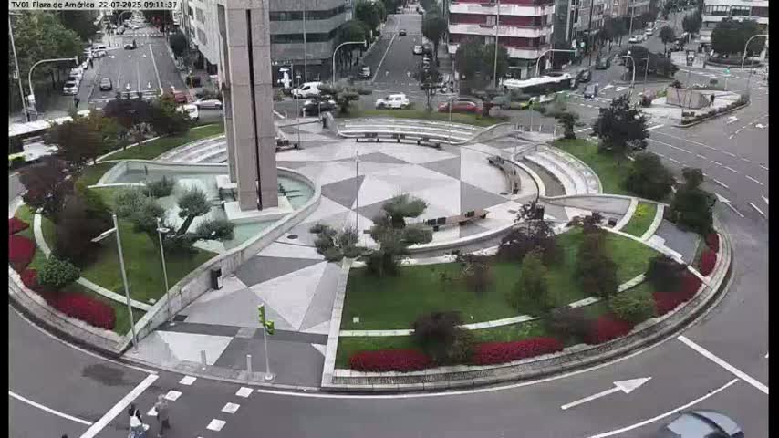 An aerial view shows a landscaped traffic circle with a geometric pattern, featuring low shrubs, small trees, and benches, under an overcast sky.