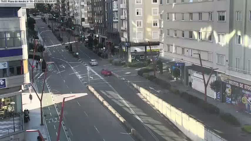 A sunny day reveals a mostly empty city street with a few cars and pedestrians, bordered by buildings and some landscaping.