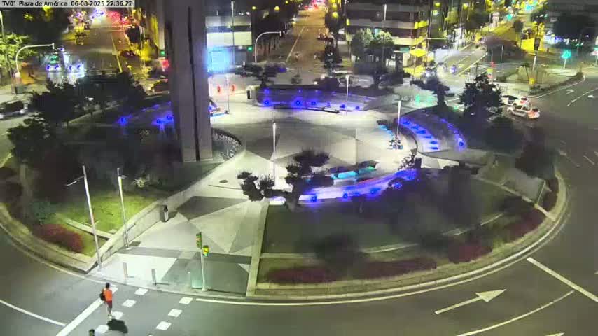 A nighttime aerial view shows a circular plaza with blue lights, landscaping, and a roadway around it, with a person walking across a crosswalk and vehicles driving on nearby streets.