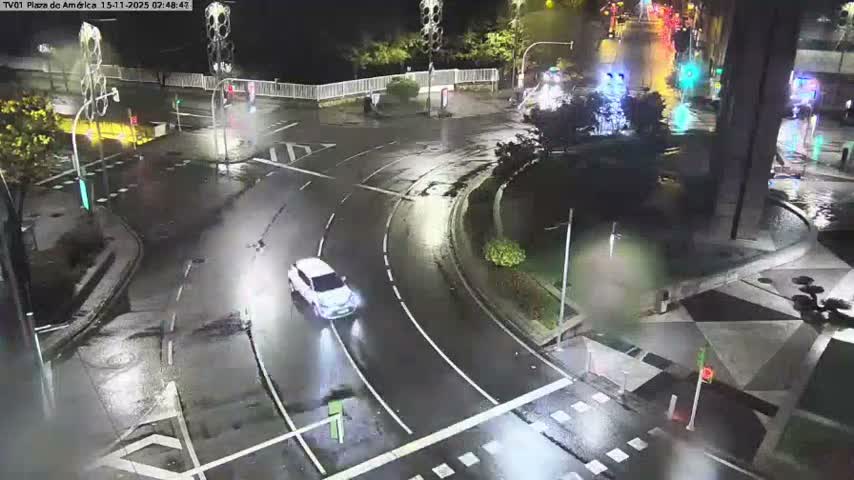 A white car drives on a wet, illuminated city street with multiple traffic lights at night, indicative of rainy weather.