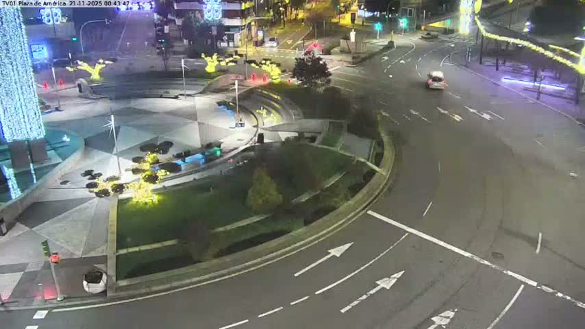 A white car drives on a wet, illuminated city street with multiple traffic lights at night, indicative of rainy weather.