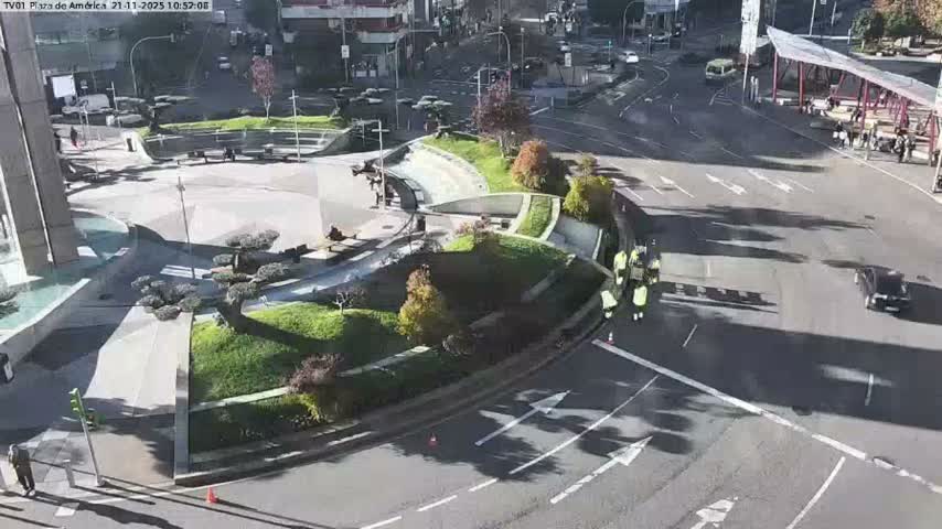 The image depicts a bustling urban plaza and intersection on a sunny day, featuring pedestrians, vehicles, green spaces, and a group of workers in high-visibility clothing.