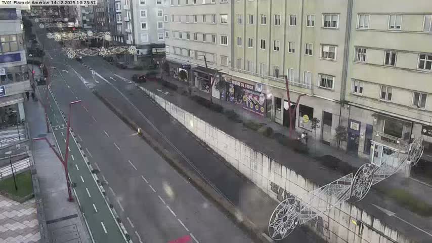A busy urban intersection is seen from an elevated perspective on an overcast day, with a cluster of cars in traffic, pedestrians on sidewalks, and a large modern building featuring a water fountain.