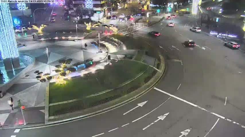 A busy urban intersection is seen from an elevated perspective on an overcast day, with a cluster of cars in traffic, pedestrians on sidewalks, and a large modern building featuring a water fountain.