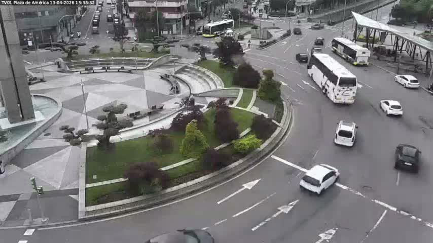 A busy urban intersection is seen from an elevated perspective on an overcast day, with a cluster of cars in traffic, pedestrians on sidewalks, and a large modern building featuring a water fountain.